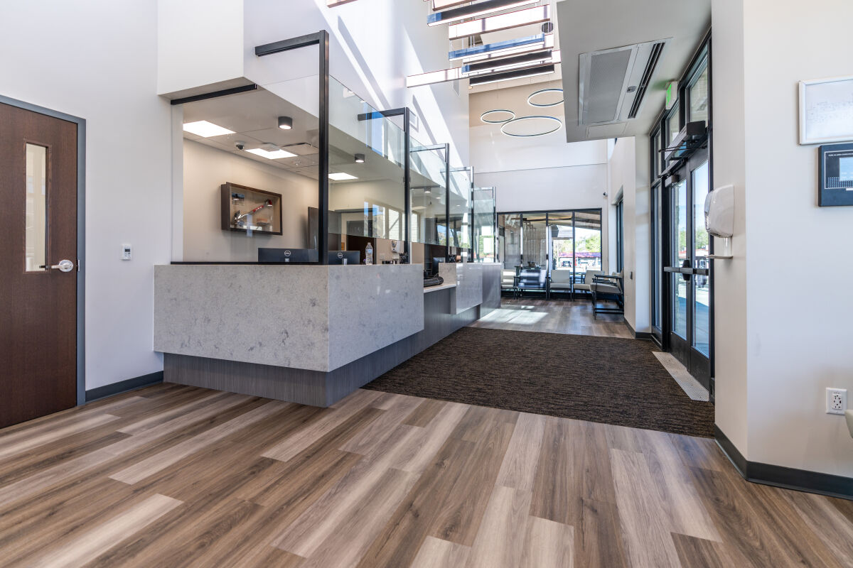 A clinic reception area with a stone‑front service desk, glass partitions, wood‑look flooring, a corridor leading to seating, and a glazed entry at the front of the space.