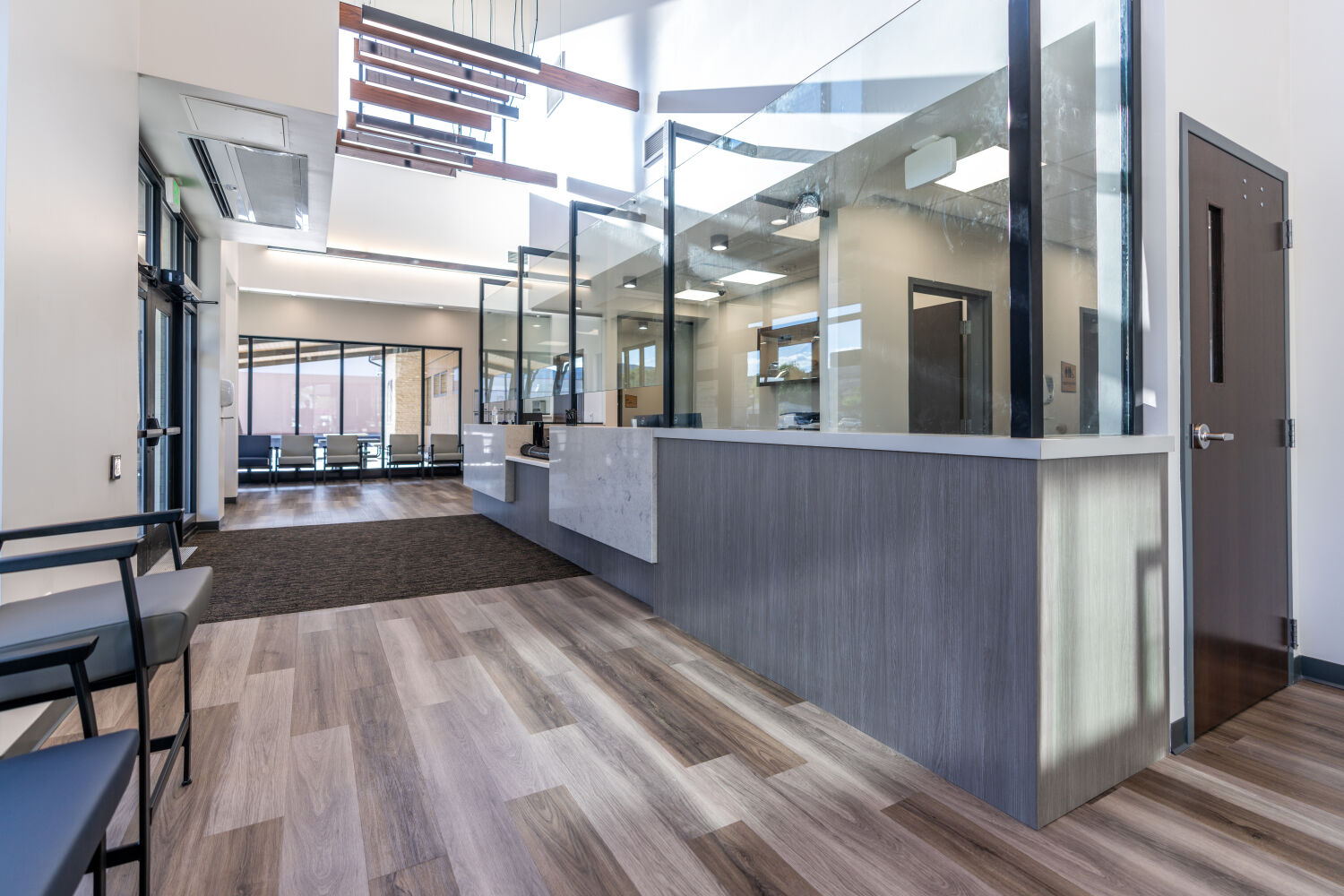 A clinic reception desk with stone and wood finishes, full‑height glass partitions, adjacent seating along the corridor, and wood‑look flooring guiding circulation through the space.