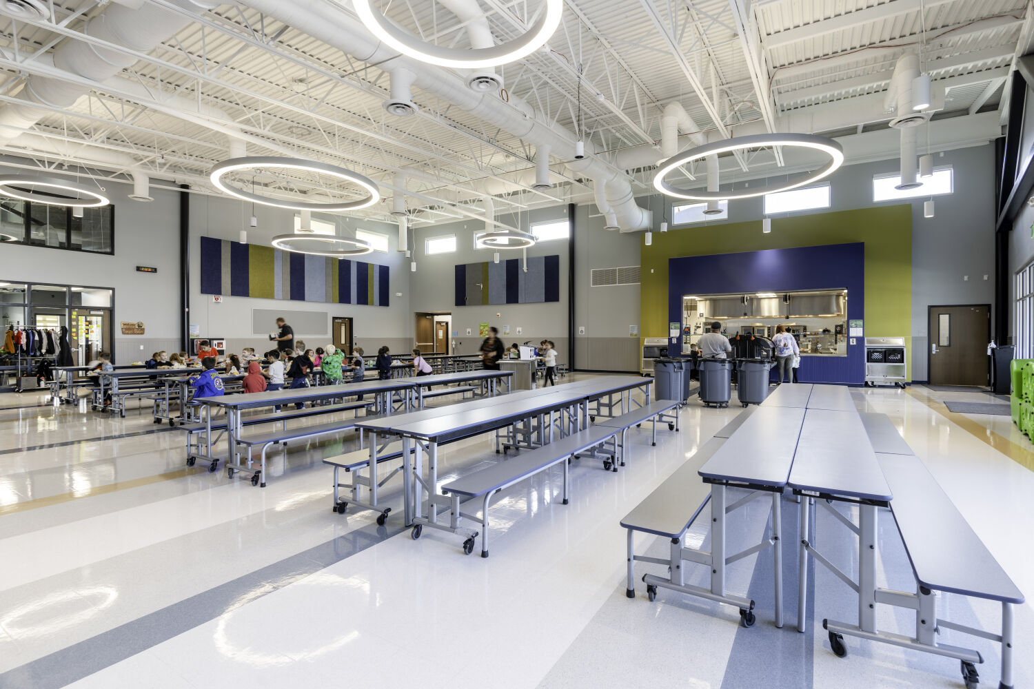 School cafeteria with long communal tables, bench seating, a food service opening, resilient flooring, acoustic wall panels, and circular suspended light fixtures.