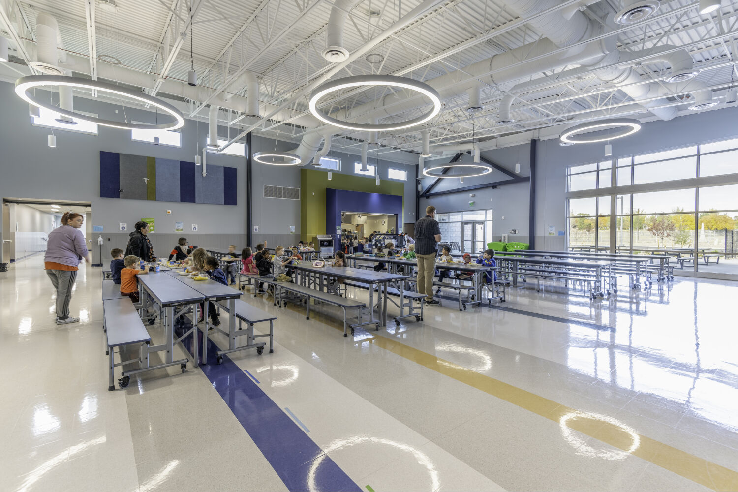 School cafeteria with long communal tables, bench seating, polished resilient flooring, large perimeter windows, and circular suspended light fixtures.