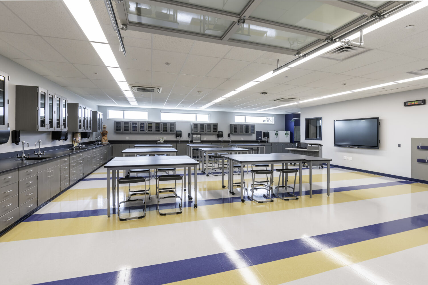 Science classroom with work tables, rolling stools, perimeter casework with sinks, upper storage cabinets, wall‑mounted display screen, and resilient flooring.