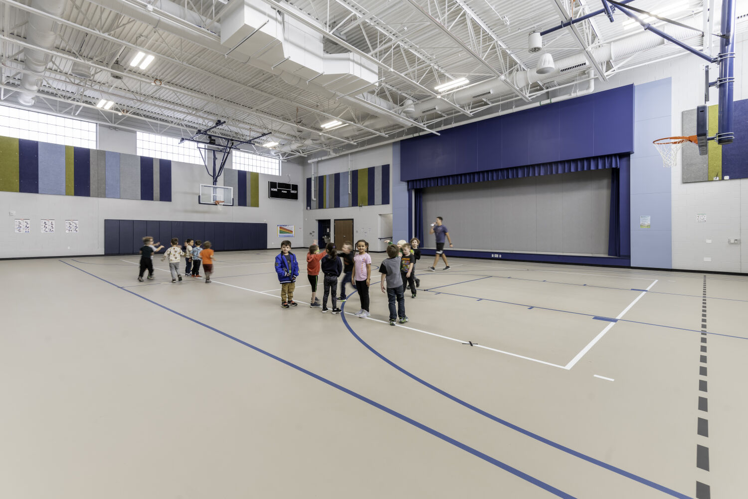 School gymnasium with a multipurpose court layout, basketball hoops, acoustical wall panels, retractable stage curtain, and high exposed ceiling structure.