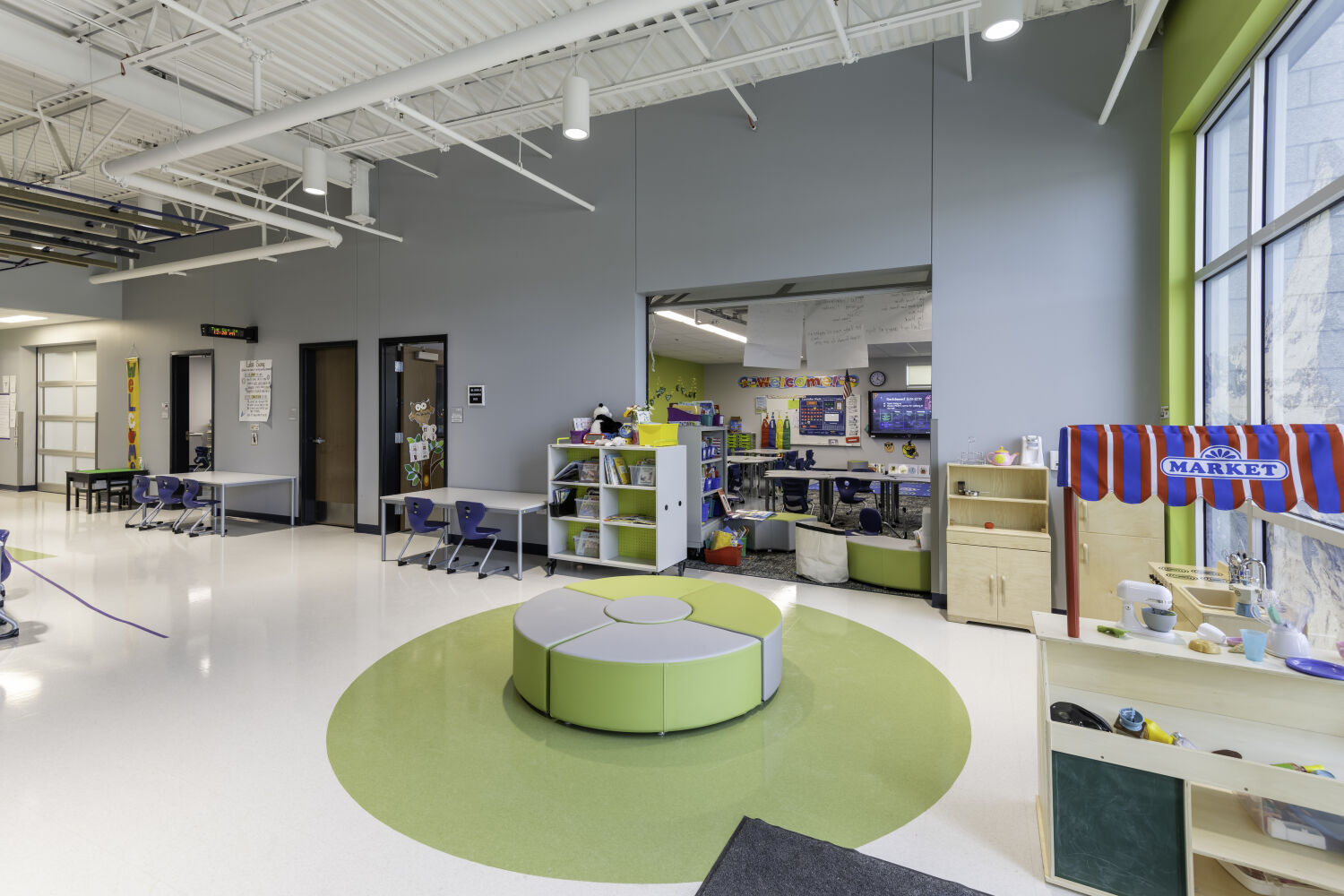 Early learning commons with modular soft seating, classroom sightlines, child‑scale furniture, materials storage, and overhead acoustic panels.