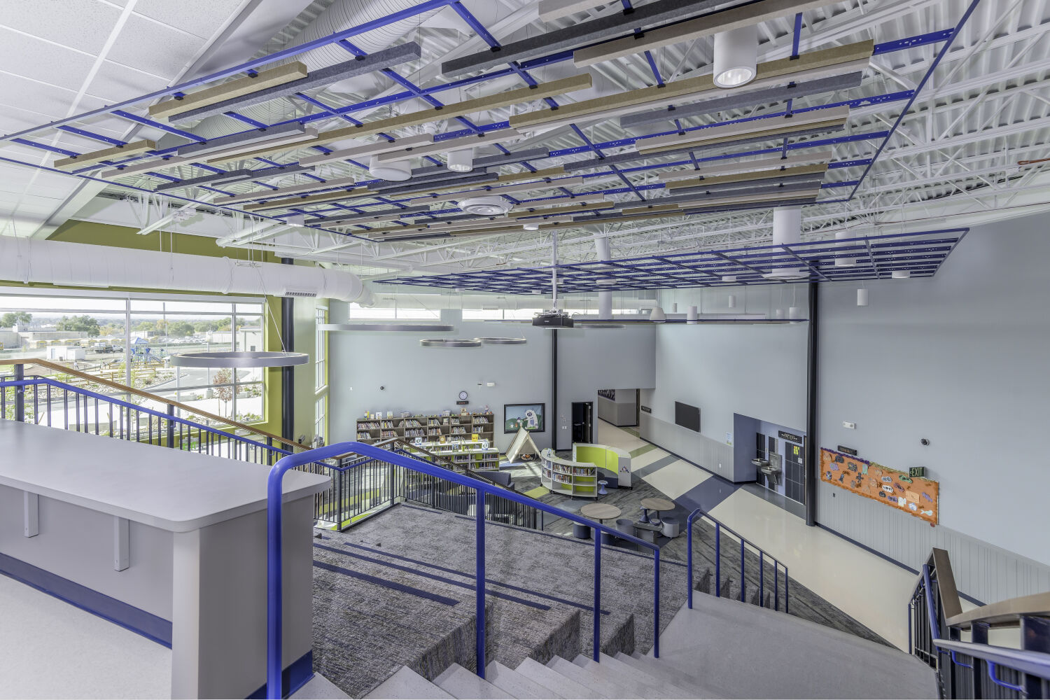 Stair overlook of a learning commons with tiered seating, central library area, open circulation paths, and suspended acoustic ceiling elements.