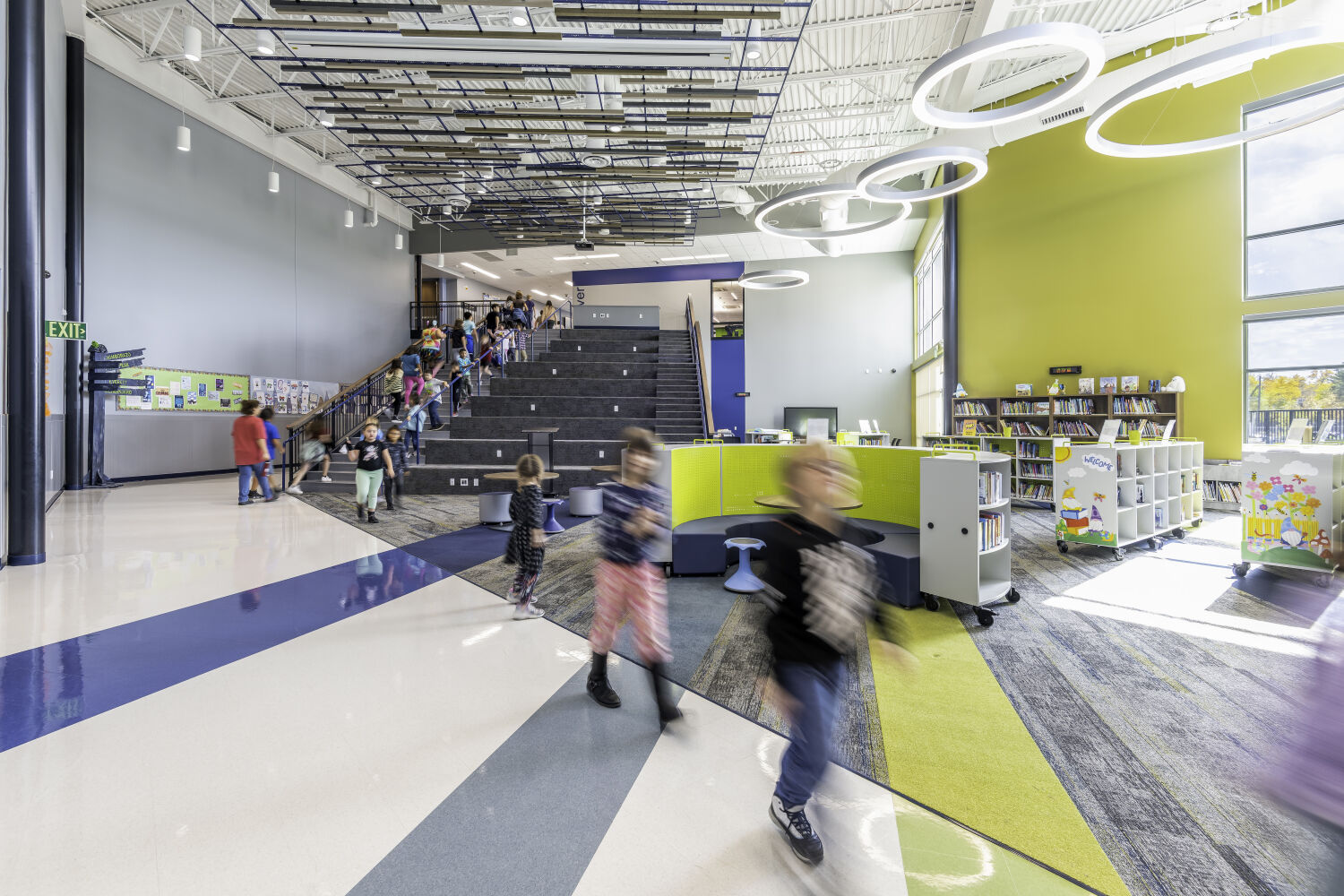 Central learning commons with a wide tiered stair, open circulation paths, mobile library shelving, soft seating areas, and suspended acoustic ceiling panels.