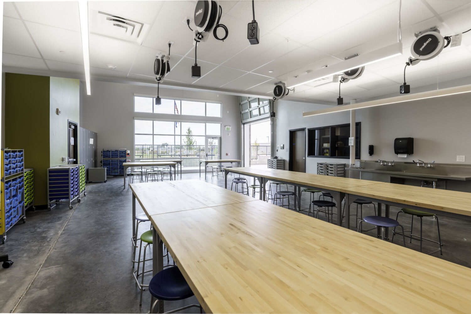 Makerspace with large worktables, stools, ceiling‑mounted power reels, materials storage carts, sinks, and a glazed overhead door.