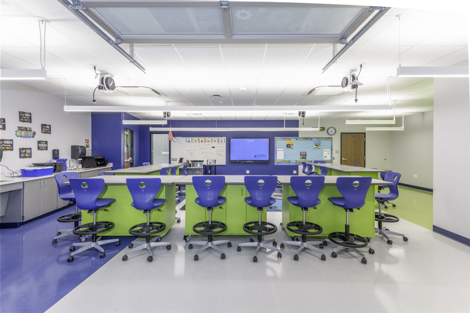 STEM classroom with lab-height tables, rolling stools, integrated technology displays, ceiling-mounted power reels, and flexible casework.