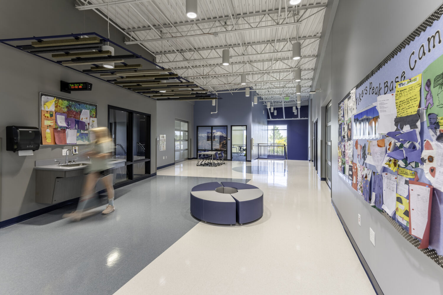 Learning corridor with a circular soft‑seating area, student work display boards, classroom glazing, a wall‑mounted sink, and suspended acoustic ceiling elements.