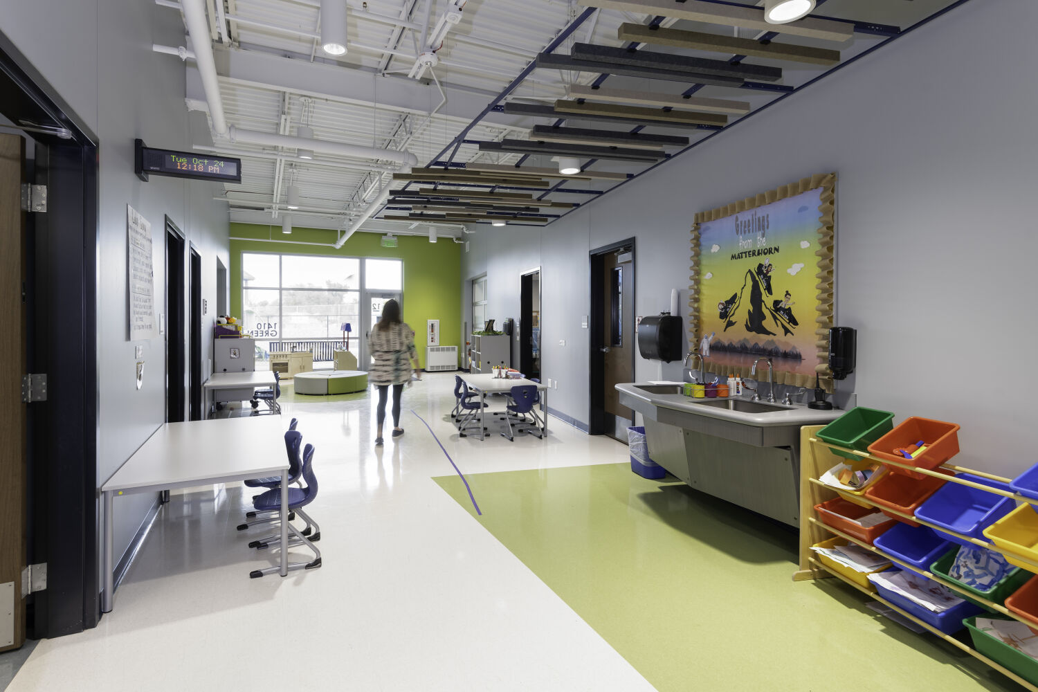 Flexible learning studio with student work tables, a sink and materials storage area, glass-front classroom doors, and acoustic ceiling panels.