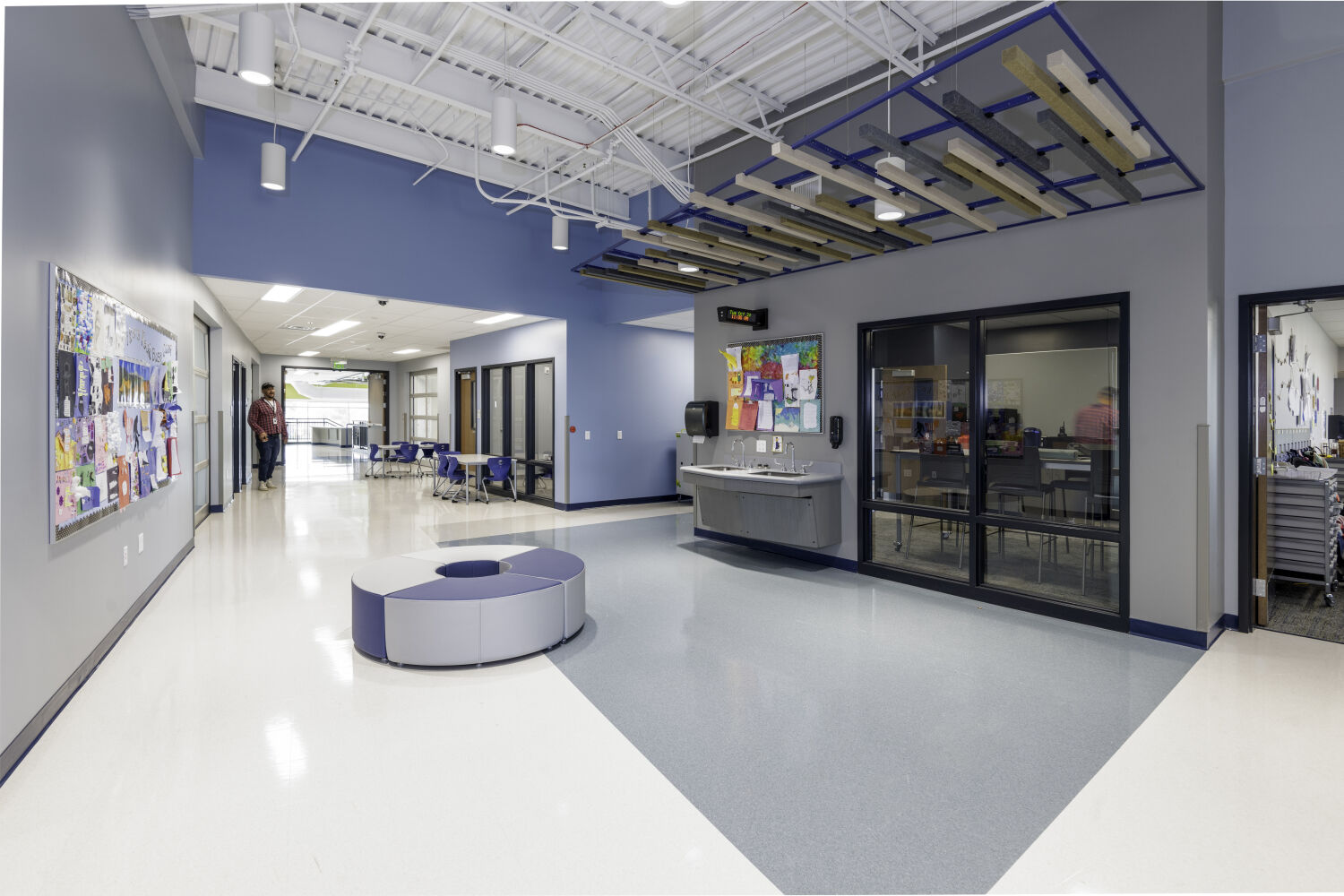 Learning corridor with circular soft seating, classroom glazing, a wall‑mounted sink, student artwork displays, and suspended acoustic ceiling elements.