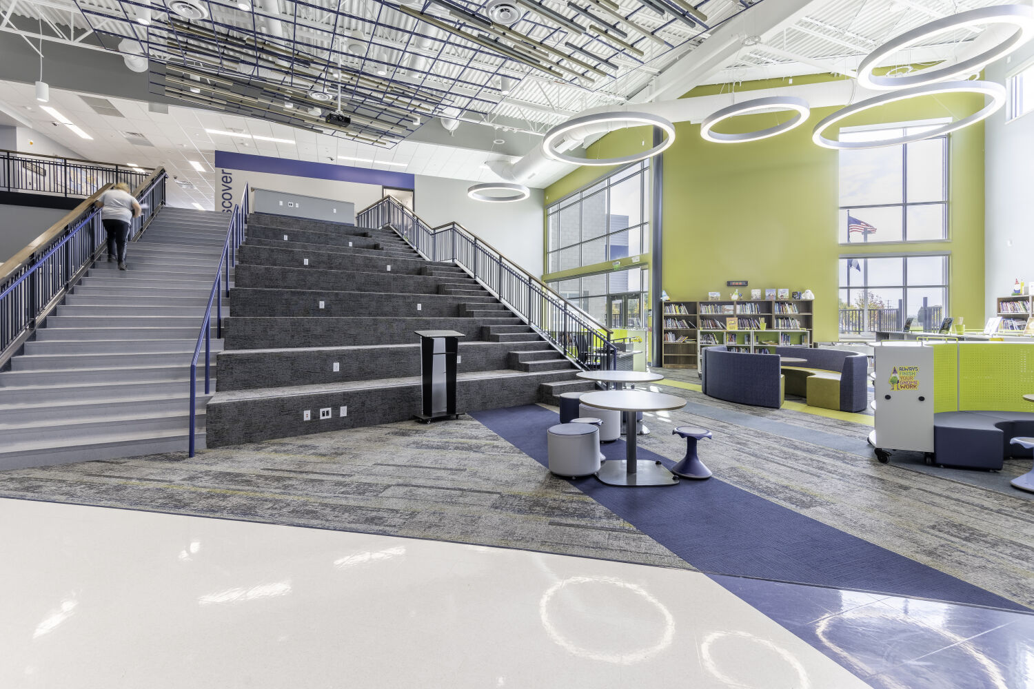 Learning commons with a wide tiered stair for seating, adjacent circulation stair, soft seating areas, mobile library shelving, and suspended acoustic ceiling elements.