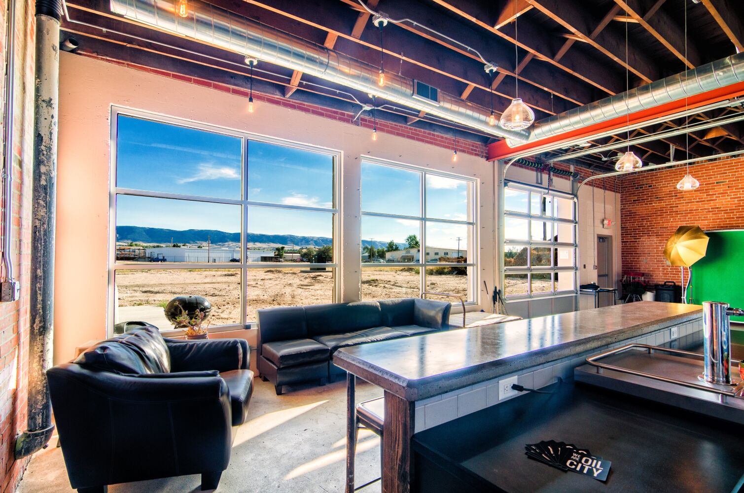 Open studio space with large industrial windows, exposed wood ceilings and ductwork, a seating area with leather couches, a long worktable, and a brick accent wall overlooking an outdoor landscape.