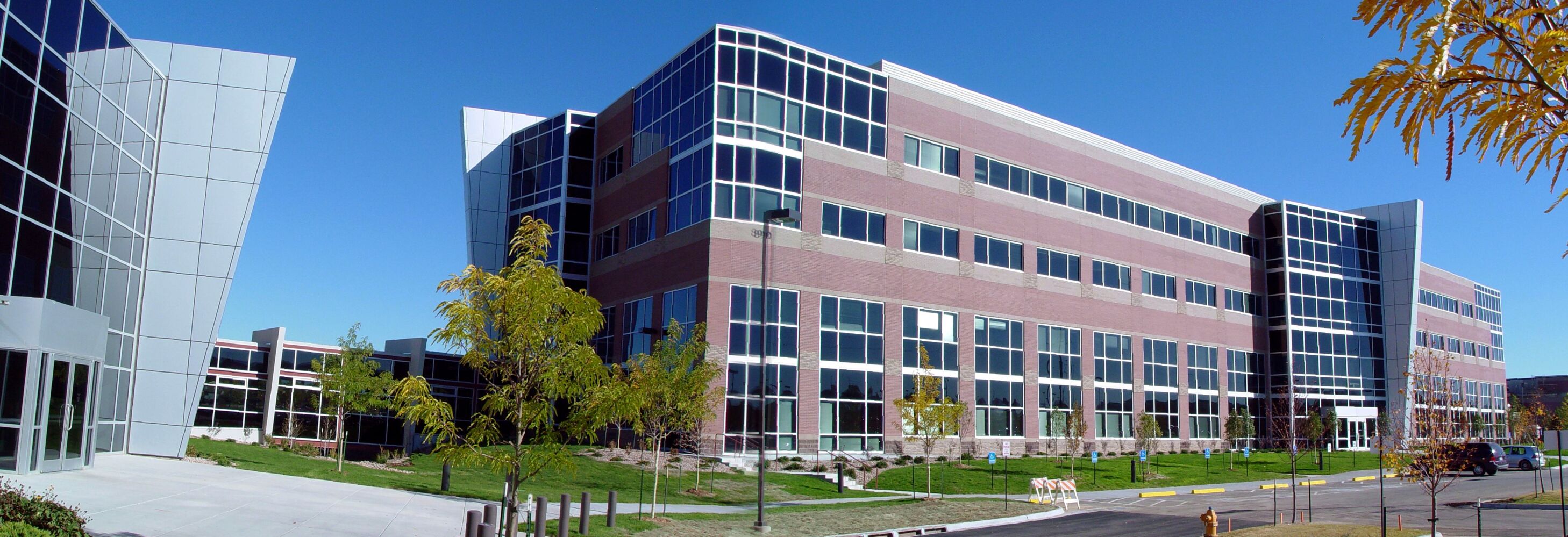 Panoramic exterior view of a modern multi‑story office building with brick and glass façades, angular glass entry volumes, landscaped lawns, young trees, and a paved roadway under a clear blue sky.