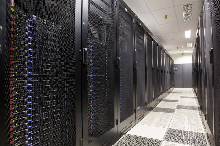 Data center aisle with rows of tall black server cabinets on both sides, illuminated overhead lighting, and a raised perforated floor for cooling airflow.