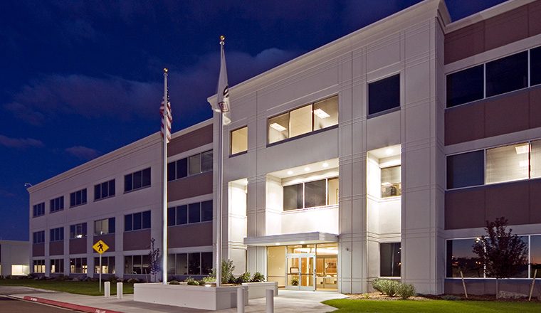 Nighttime exterior view of a three‑story office building with illuminated entry canopy, large windows, and two flagpoles near the front walkway.