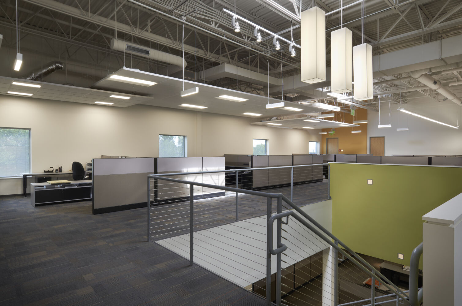 Open office area on an upper floor with cubicles, exposed structural ceiling, suspended light fixtures, carpet tile flooring, and a metal stair railing overlooking a lower level.