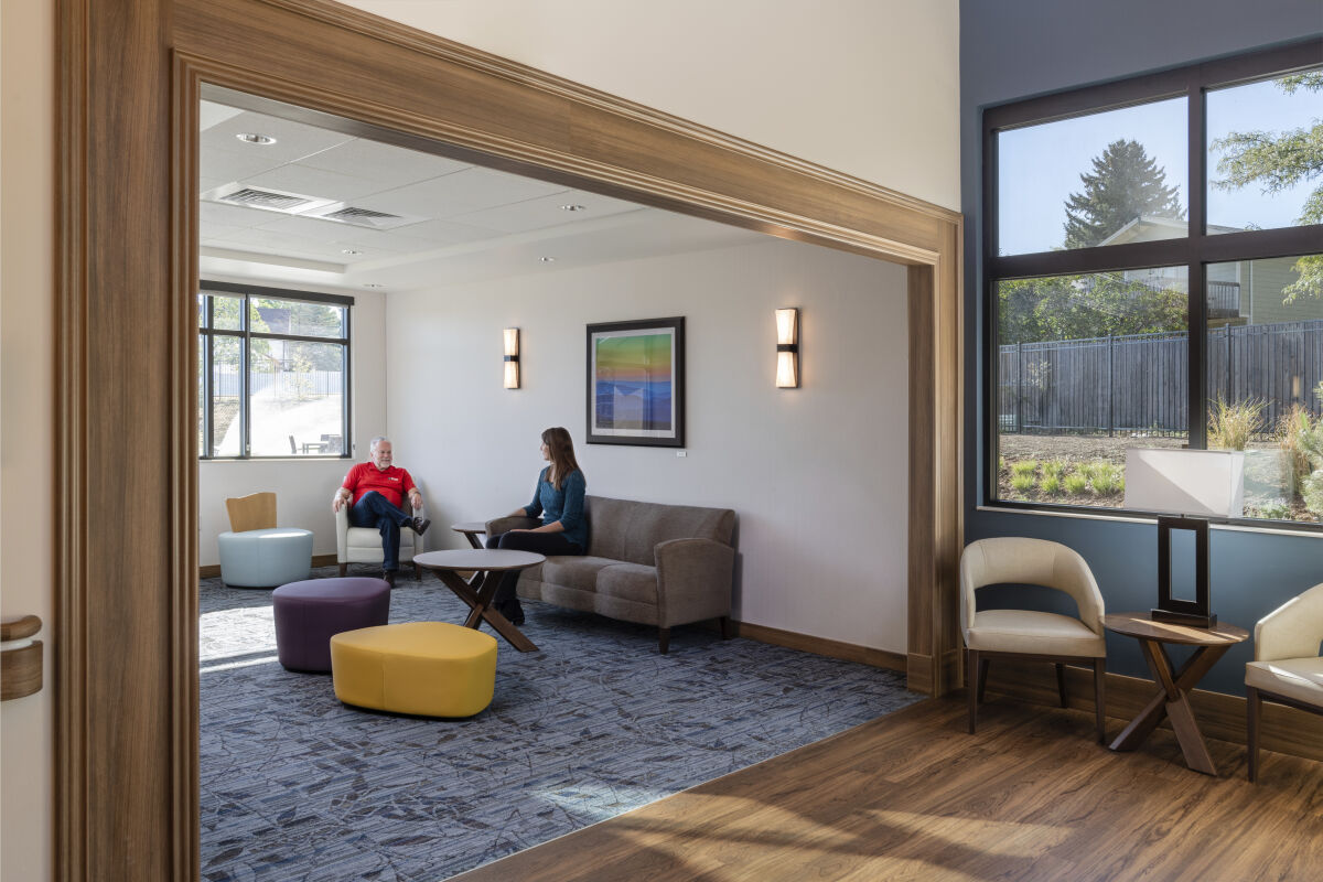 A family lounge with upholstered seating, a central coffee table, accent stools, framed artwork, wall‑mounted light fixtures, wood trim, carpeted flooring, and large windows overlooking a landscaped area.