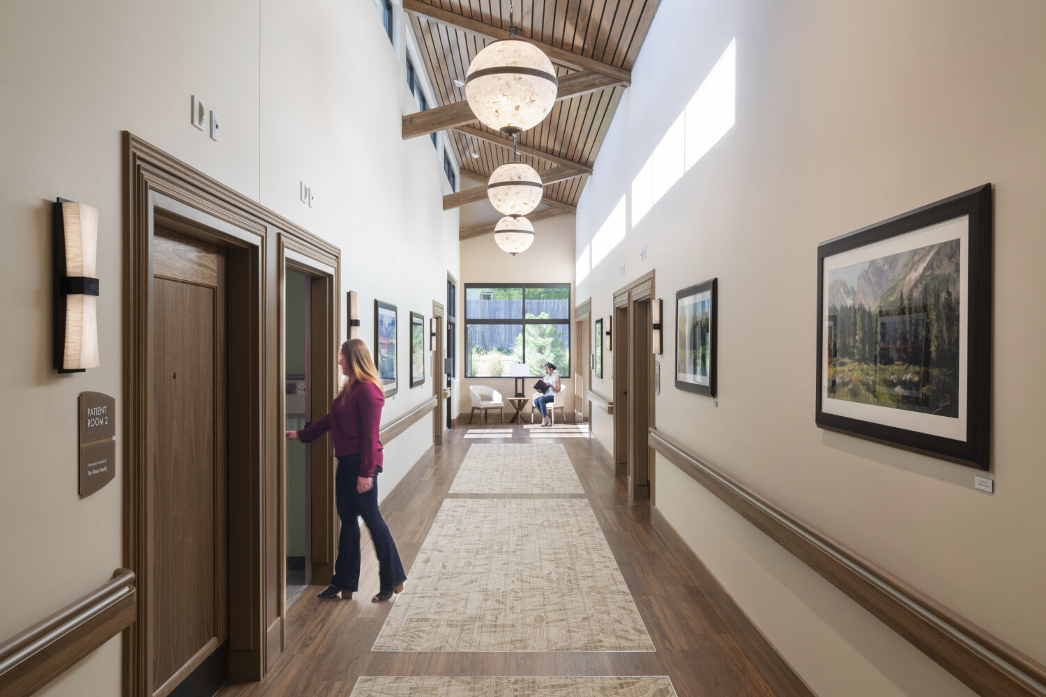 A patient room corridor with wood doors, framed artwork, wall sconces, lantern‑style pendant lights, clerestory windows, and a patterned runner defining the circulation path.