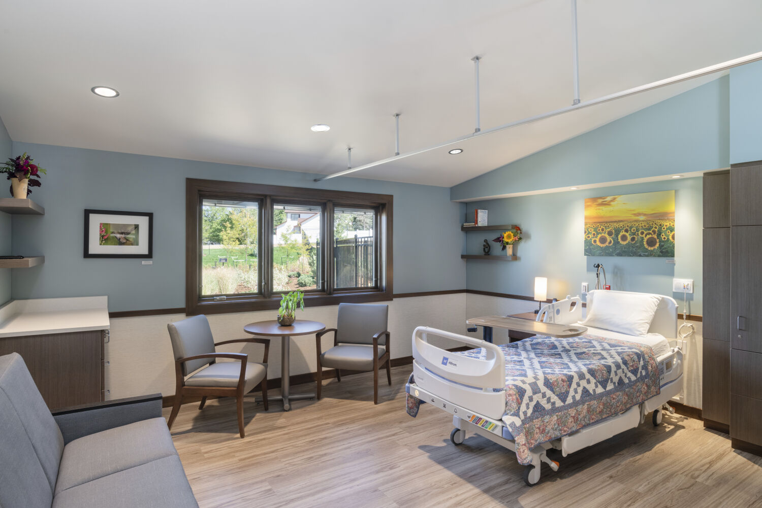 A patient room with a hospital bed, bedside lighting and controls, upholstered guest seating, a small table and chairs near the window, built‑in cabinetry, wood‑look flooring, and calming artwork on the walls.