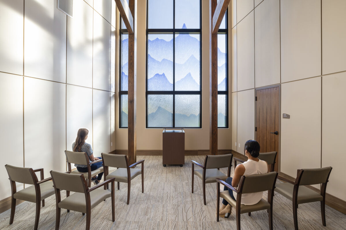 A quiet reflection room with arranged seating, tall wood columns, a central lectern, soft wall panels, carpeted flooring, and a large decorative window featuring layered landscape imagery.