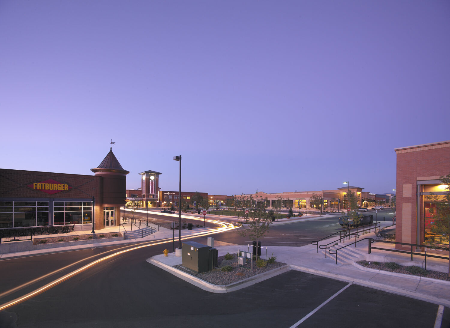 Dusk view of a town center with illuminated storefronts, a curved roadway, and light trails from passing vehicles, with the Fatburger building and a distant clock tower visible.