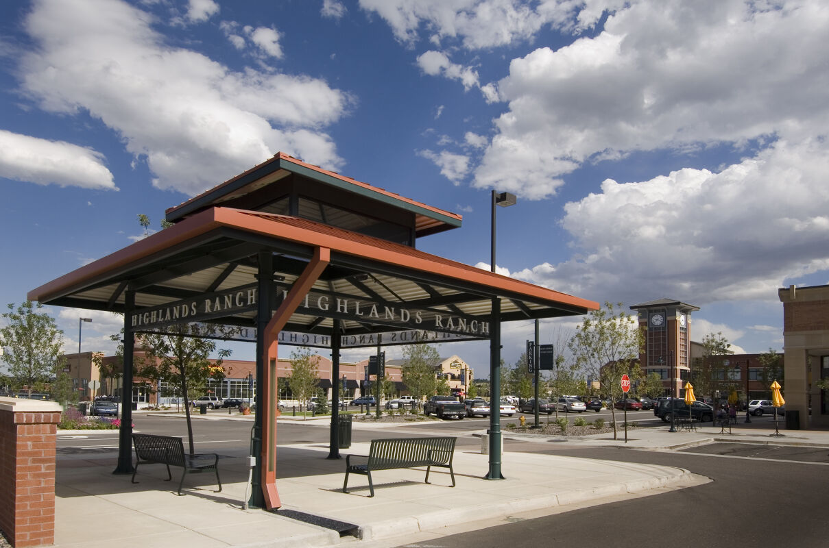 Open-air pavilion with a metal roof, benches, and surrounding landscaping, set within a town center area with storefront buildings and a distant clock tower.