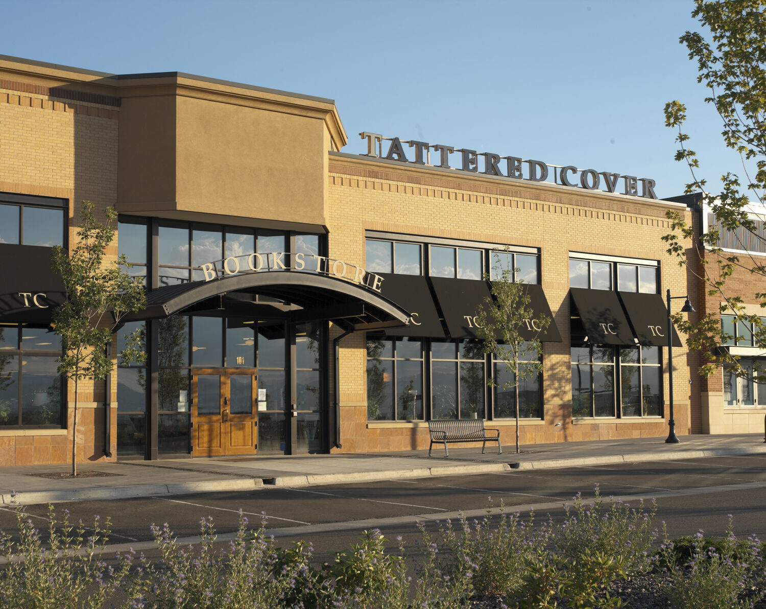 Exterior view of the Tattered Cover bookstore with tan brick façades, black awnings, a curved entrance canopy labeled “BOOKSTORE,” and street‑side landscaping.