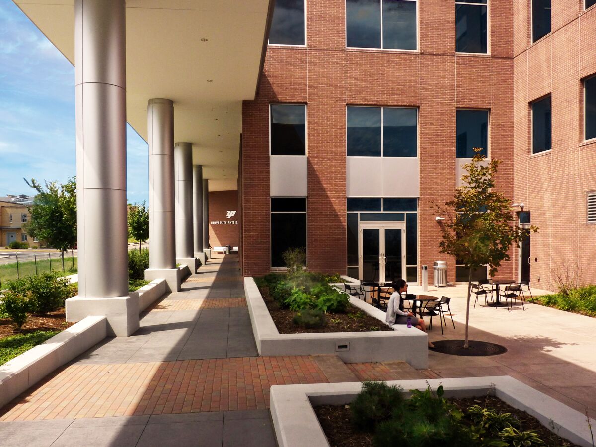 Covered walkway alongside a multi‑story brick office building with planters, young trees, and outdoor seating areas along a landscaped pedestrian path.