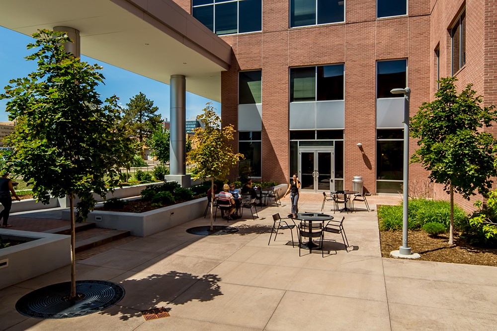 Outdoor courtyard with seating areas, young trees, planters, and a covered walkway adjacent to a multi‑story brick office building.