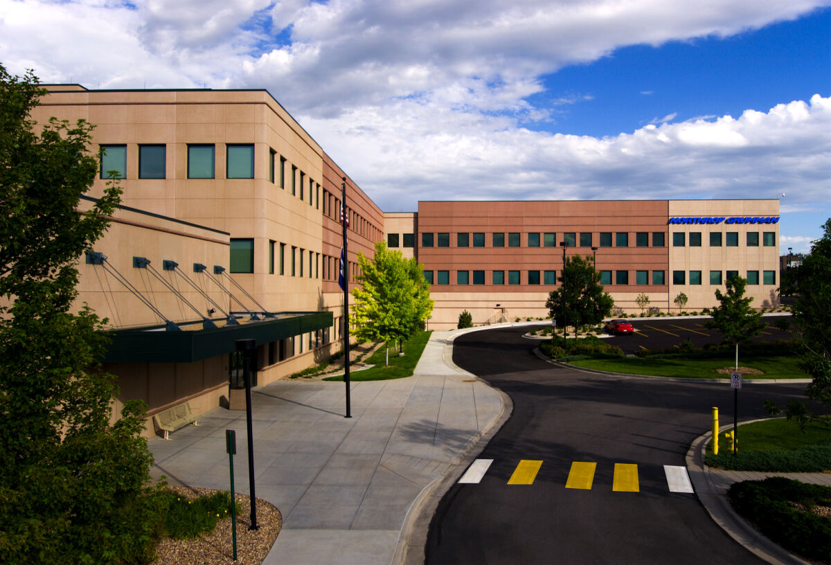 Daytime exterior view of a multi‑story office building with a glass canopy entry, landscaped walkways, and a curved roadway leading to the Northrop Grumman facility.