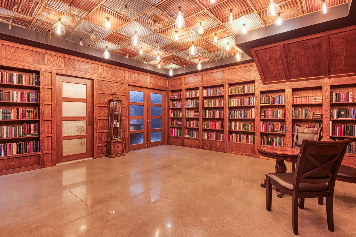 Library-style entrance to Aiden Sinclair’s Underground featuring built-in bookshelves, wood-paneled walls, frosted glass doors, a polished concrete floor, and overhead lighting with exposed bulbs.