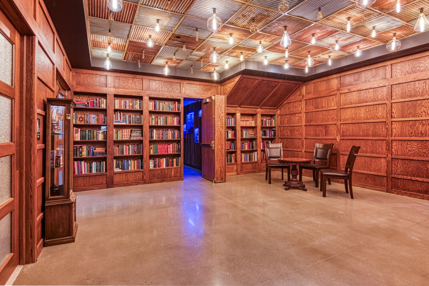 Library-style room inside Aiden Sinclair’s Underground featuring wood-paneled walls, built-in bookshelves filled with books, a polished concrete floor, and an overhead grid of exposed lightbulbs.