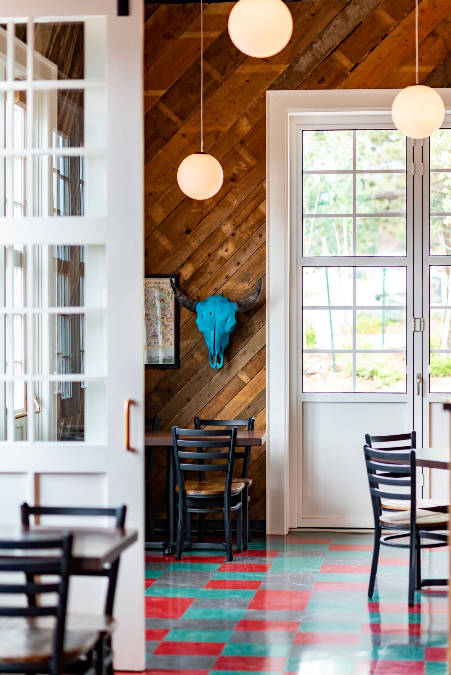 Interior view of The Post restaurant showing a diagonal wood-plank accent wall with a blue decorative skull, large windows, hanging globe lights, and red-and-green patterned flooring.