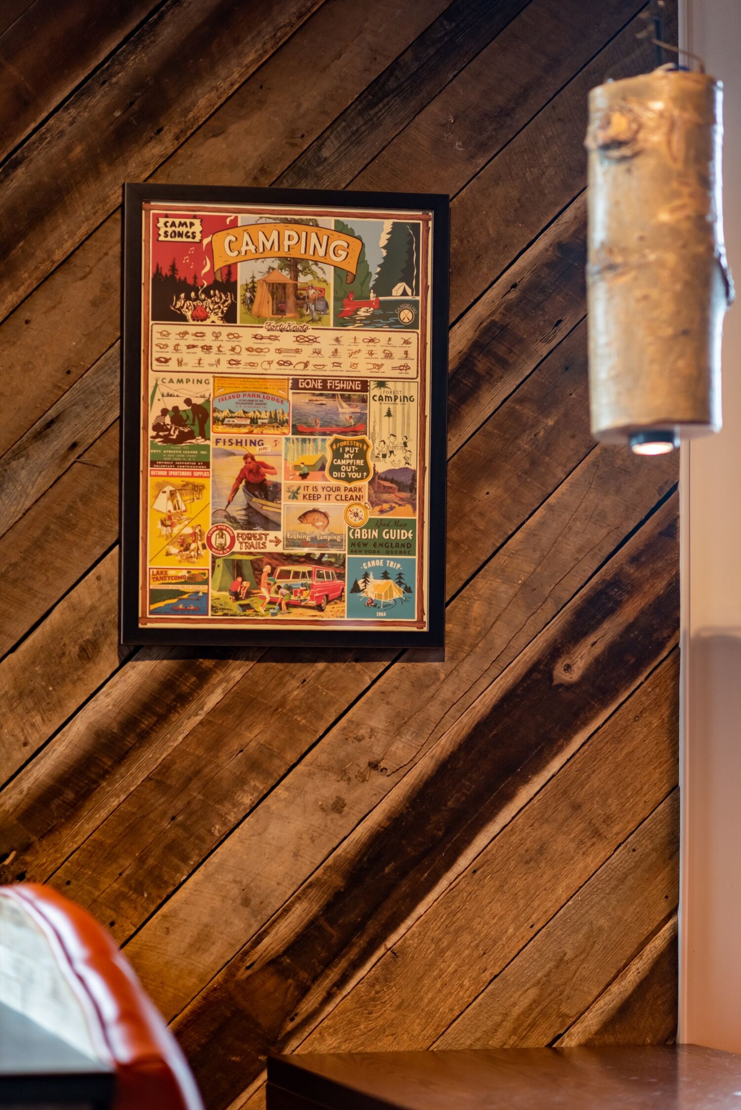 Framed vintage camping poster displayed on a diagonal wood-plank wall with a cylindrical hanging light fixture nearby.