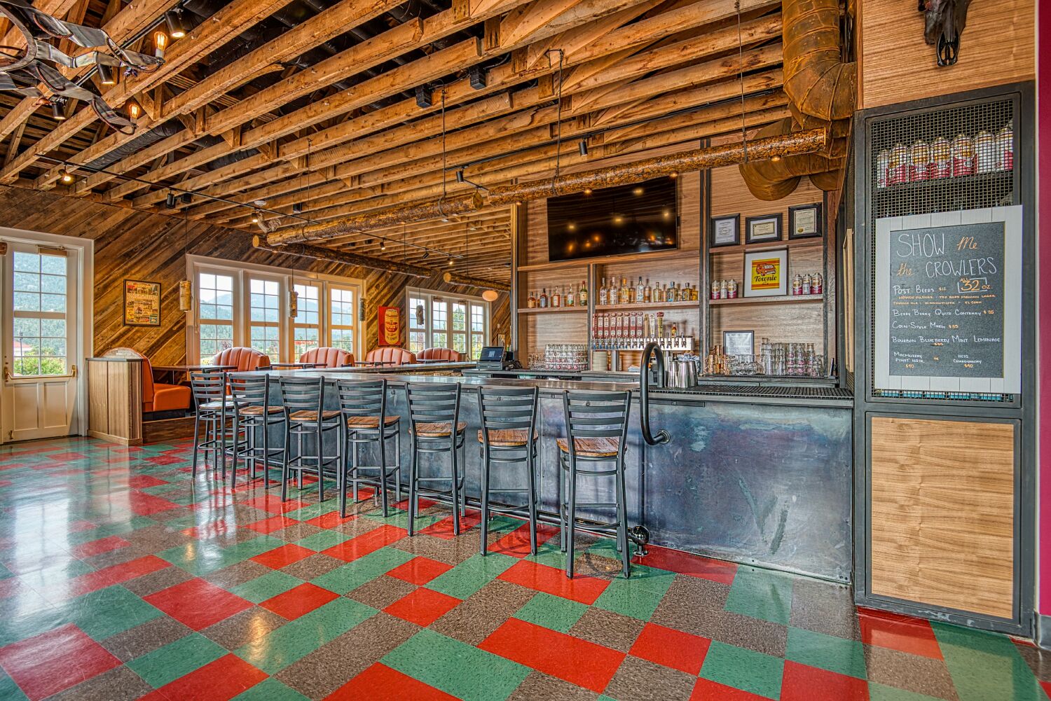 Bar interior at The Post restaurant featuring a long counter with metal barstools, exposed wood ceiling structure, built‑in shelving with bottles and glassware, and red‑and‑green patterned flooring.