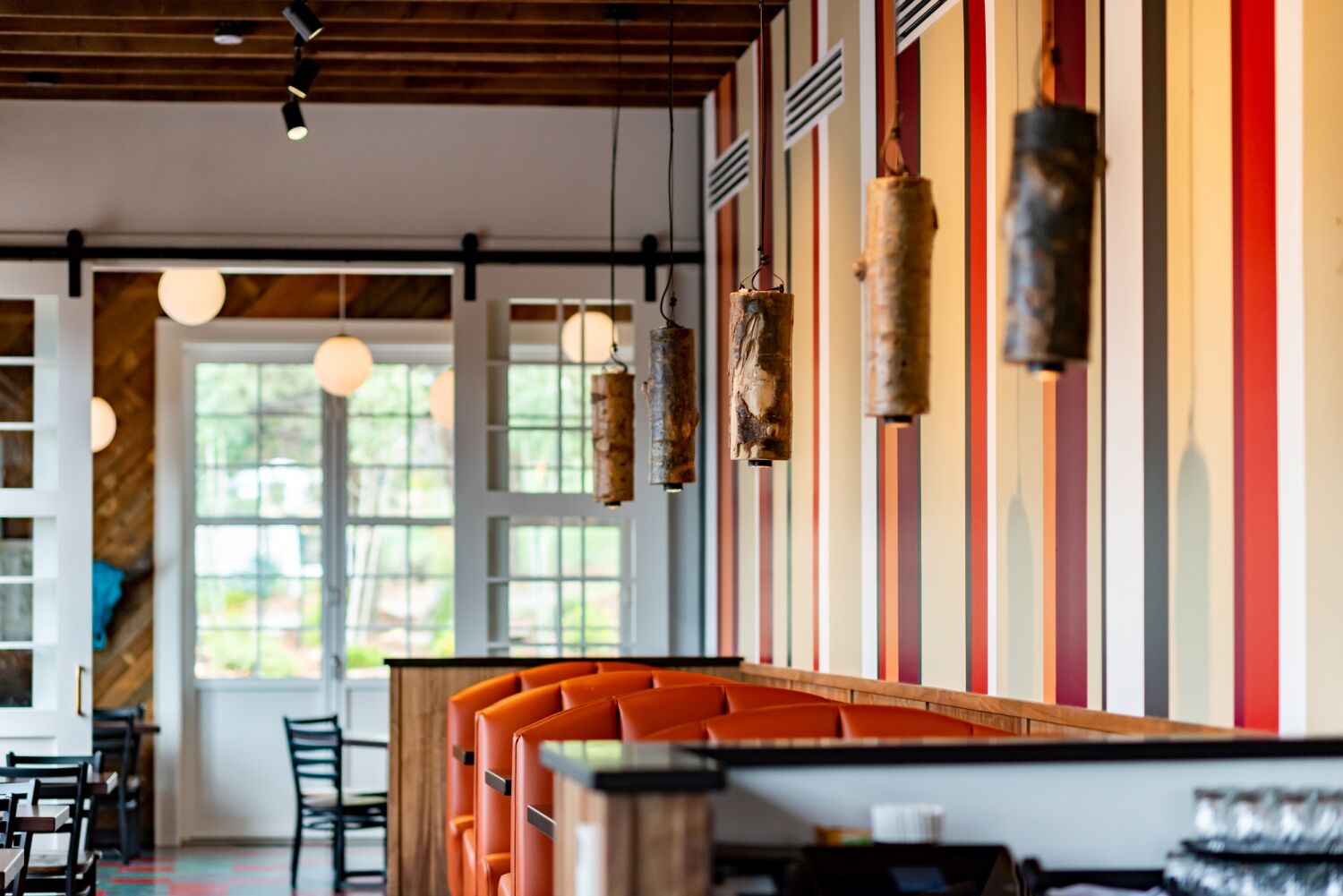 Interior view of The Post restaurant featuring cylindrical hanging light fixtures, orange booth seating, and a striped accent wall with large windows in the background.