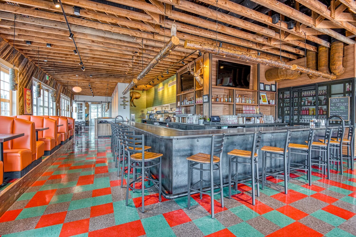Interior of The Post restaurant at the Stanley Carriage House featuring an open bar, exposed wood ceiling, orange booths, and patterned red‑and‑teal flooring.