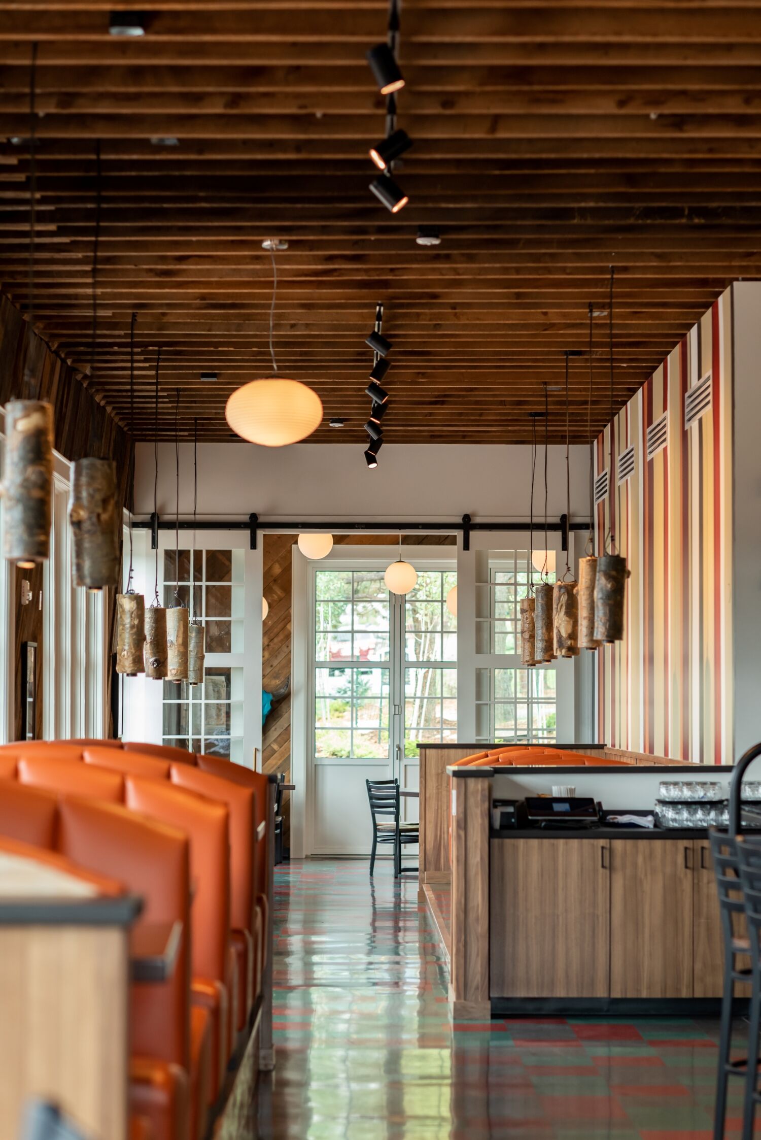 Interior view of The Post restaurant with orange booth seating, wood‑paneled ceiling structure, hanging cylindrical light fixtures, striped accent wall, and a host station near large windows.