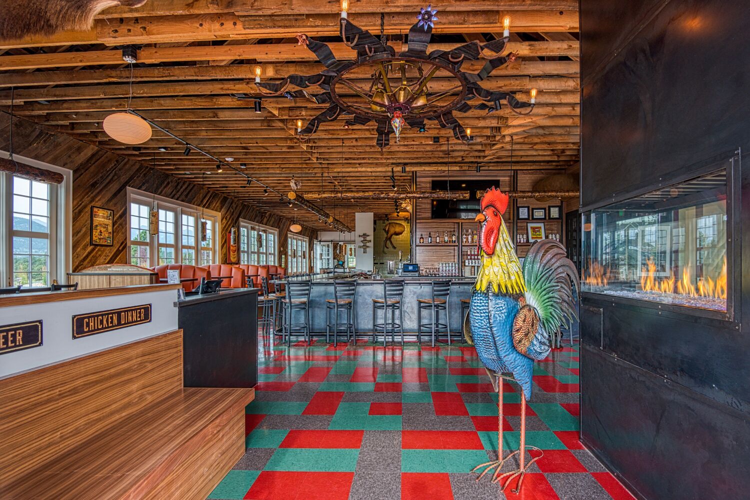 Entrance view of The Post restaurant showing a colorful rooster sculpture, double‑sided fireplace, exposed wood ceiling, bar seating, and patterned red‑and‑teal flooring.
