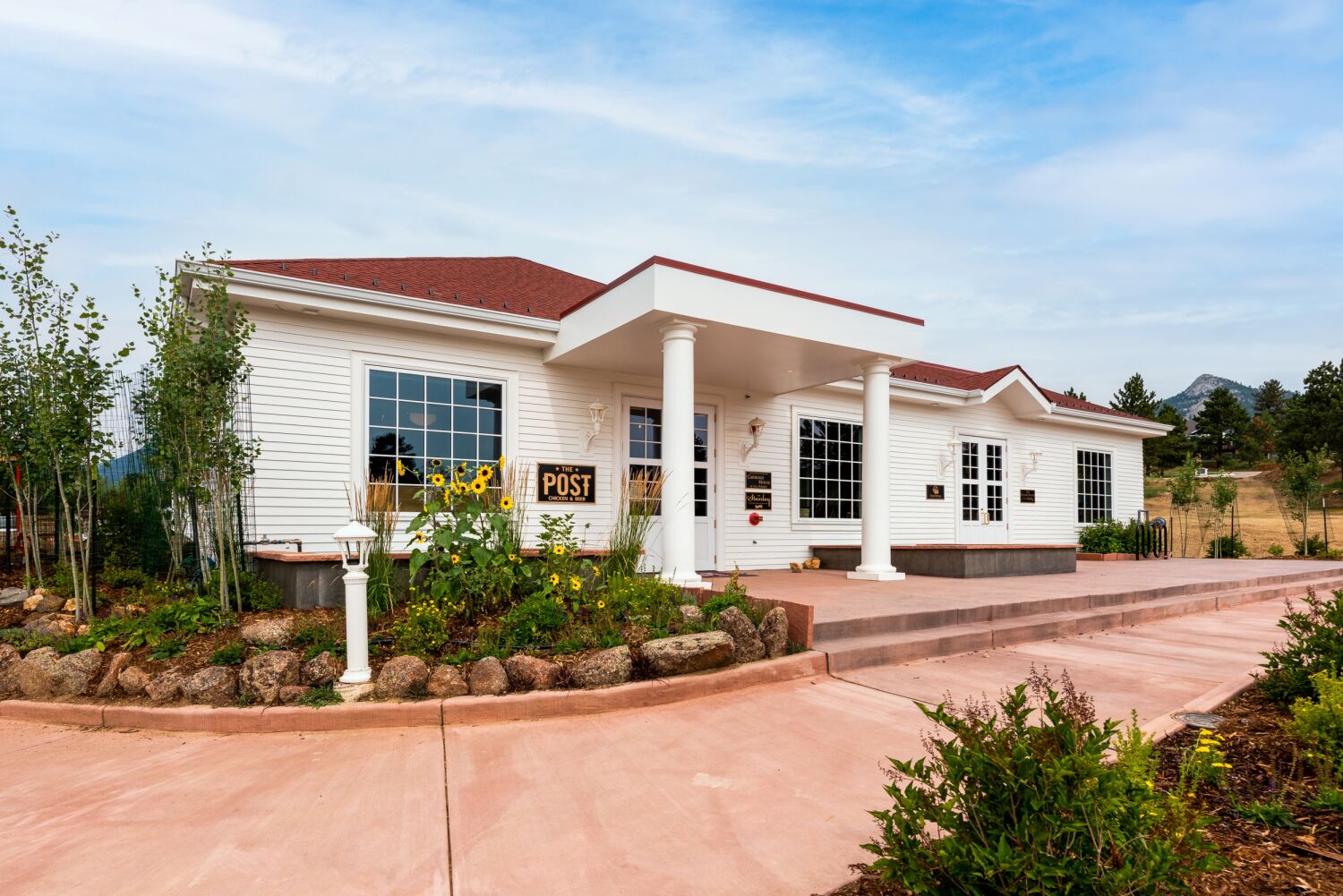 Restored white carriage house with columns, landscaped garden beds, and mountain backdrop under a blue sky.