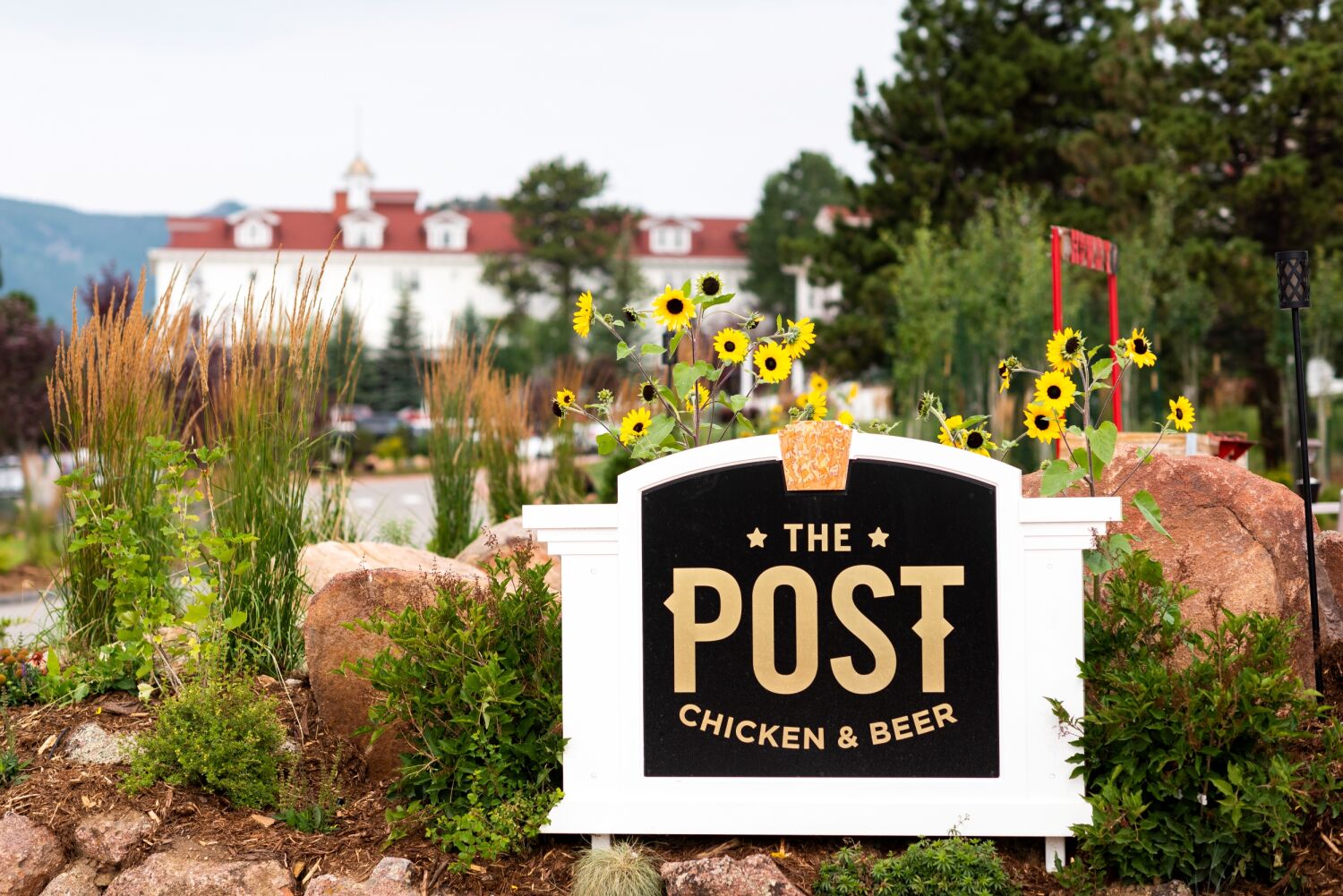Exterior sign for The Post restaurant reading ‘THE POST CHICKEN & BEER,’ surrounded by rocks, greenery, and yellow flowers with The Stanle Hotel visible in the background.