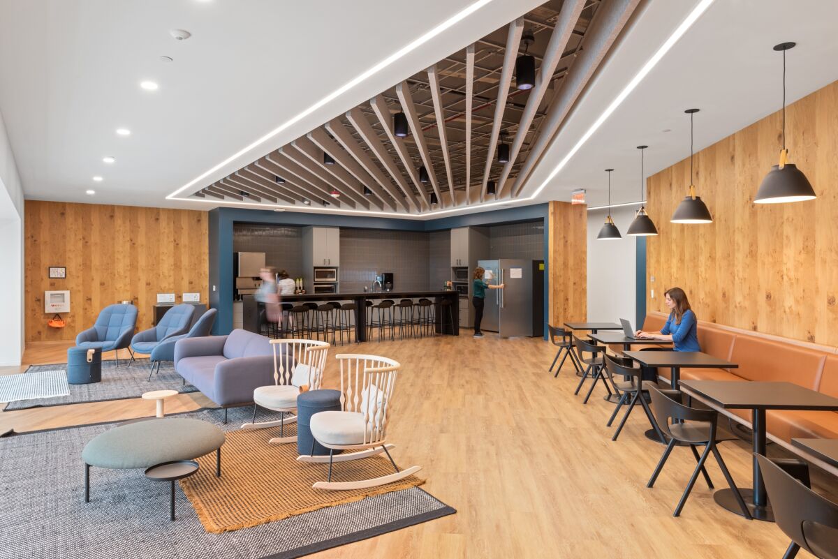 Breakroom café space in a Medtronic workplace featuring lounge seating, booth tables, pendant lighting, wood wall paneling, a slatted ceiling detail, and an open kitchenette with bar‑height seating.