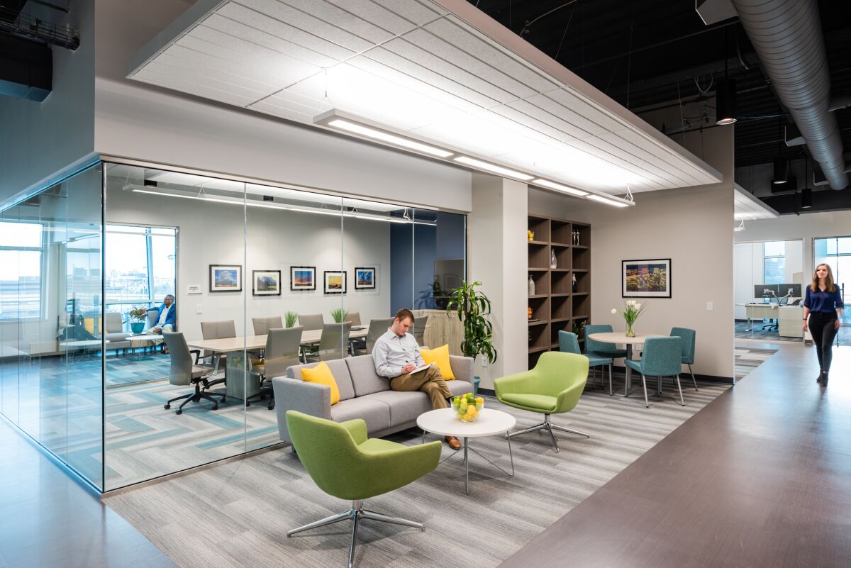 Collaboration area in the Caring for Colorado Denver office featuring a glass-front conference room, modern lounge seating with colorful pillows, built-in shelving, and overhead linear lighting.