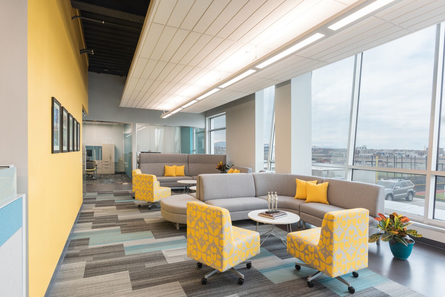 Lounge seating area in the Caring for Colorado Denver office featuring curved gray sofas with yellow accent pillows, patterned yellow chairs, round white tables, a bright yellow feature wall with framed artwork, and large windows.