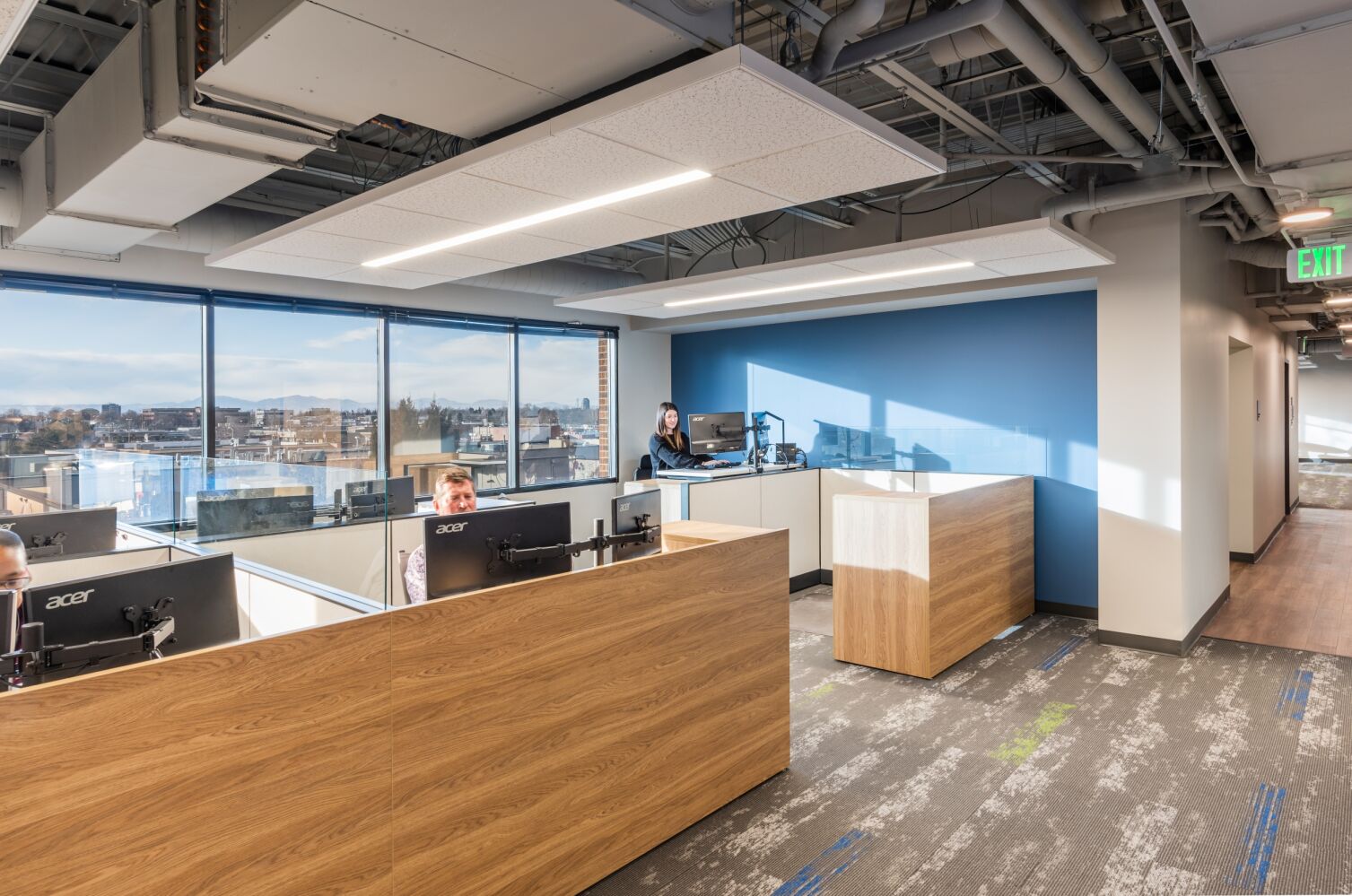 Open workstation area in the STRIDE Administration Office featuring wood‑accented desks with multiple monitors, a blue accent wall, large windows with city views, exposed ceiling structure, and patterned carpet flooring.