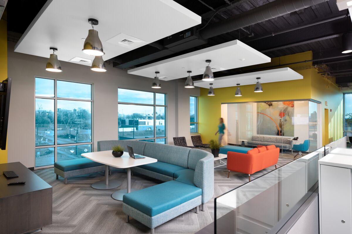 Collaboration lounge at the All Copy Headquarters featuring modular blue sectional seating around round tables, an orange sofa in a glass‑front meeting space, yellow accent walls, suspended acoustic ceiling panels, and large windows overlooking the surroun