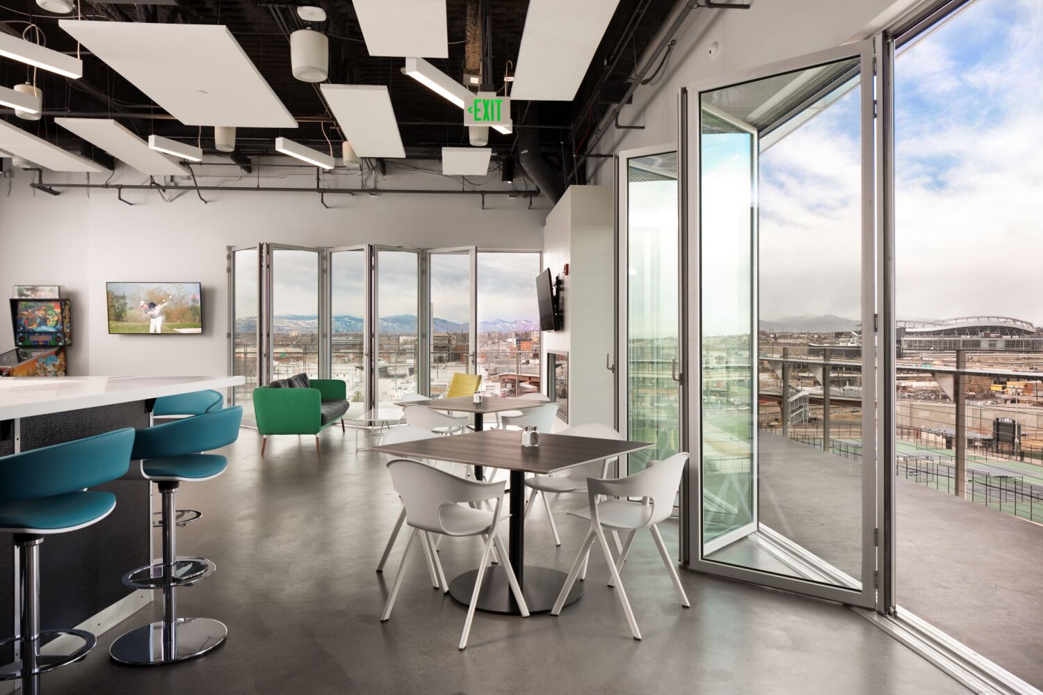 Indoor–outdoor lounge at the All Copy Headquarters featuring bar-height seating, round café tables with white chairs, large folding glass doors opening to a terrace, suspended acoustic ceiling panels, and expansive city and mountain views through floor-to-