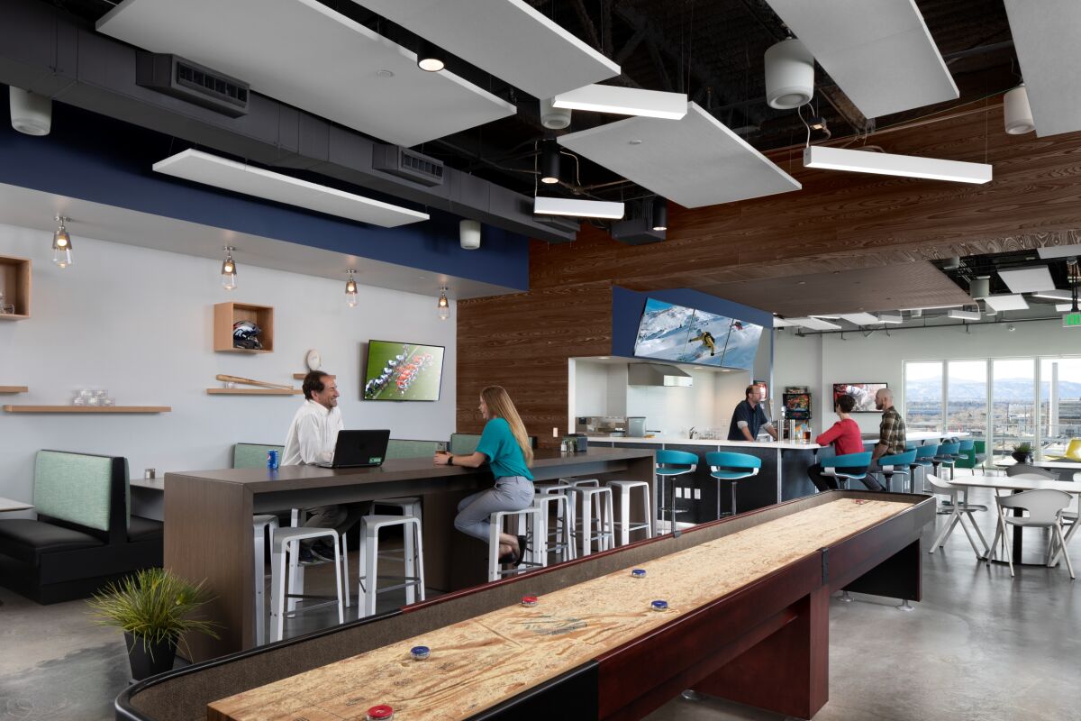Social hub at the All Copy Headquarters featuring a long café counter with bar stools, wood‑slat accent walls, suspended acoustic ceiling panels, booth seating, televisions mounted on the walls, pendant lighting, and a shuffleboard table in the foreground.