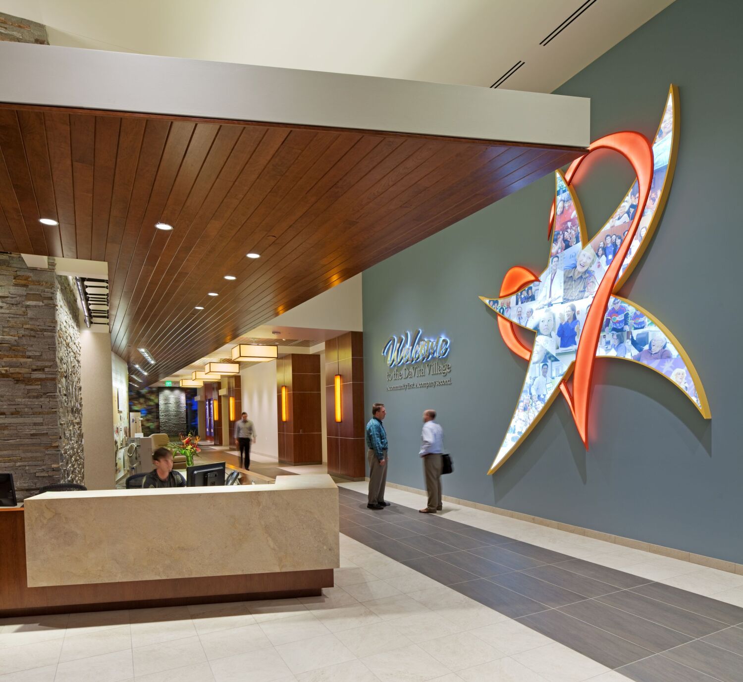 Lobby reception area at the DaVita Headquarters featuring a stone-accented desk, a wood-slat ceiling installation, a large sculptural star-shaped feature wall with integrated imagery, pendant lighting, and a tiled walkway leading into the interior corridor