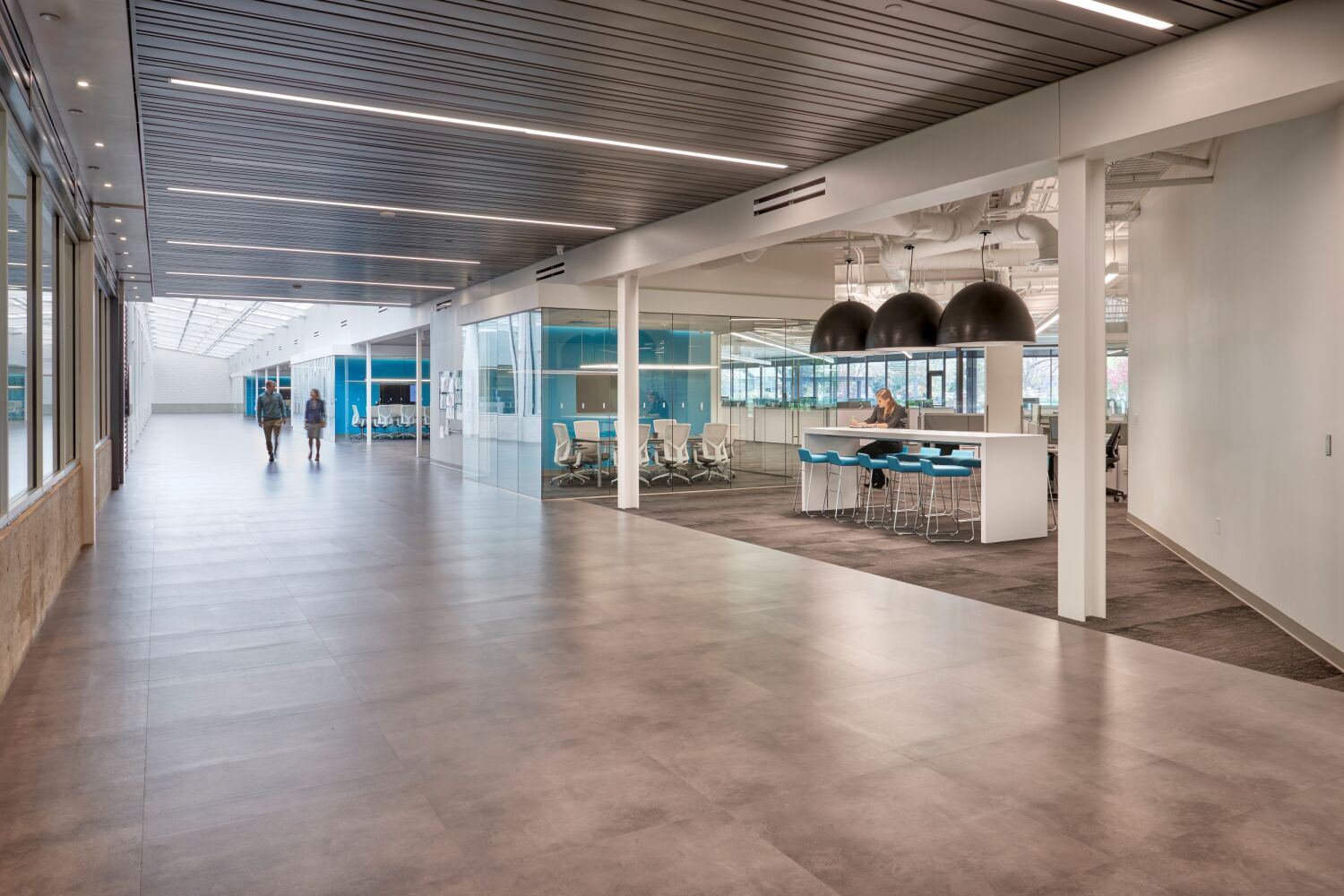 Woodward Drake corridor with a glass‑walled conference room, open collaboration area with high‑top seating, and linear ceiling lighting extending toward a skylit atrium.